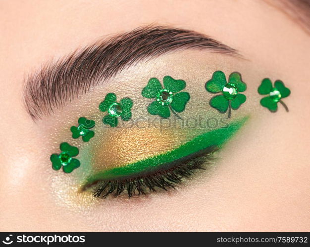 Conceptual photo of St. Patrick&rsquo;s Day. The eye of the young beautiful woman with bright green shadows and expressive eyebrows. Shamrock patterns. Holiday makeup