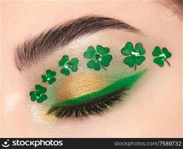 Conceptual photo of St. Patrick&rsquo;s Day. The eye of the young beautiful woman with bright green shadows and expressive eyebrows. Shamrock patterns. Holiday makeup