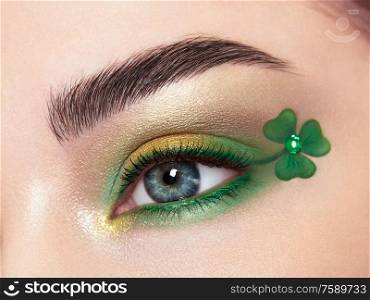 Conceptual photo of St. Patrick&rsquo;s Day. The eye of the young beautiful woman with bright green shadows and expressive eyebrows. Shamrock patterns. Holiday makeup