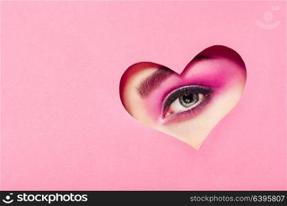 Conceptual photo of Valentine&rsquo;s day. Eye of Girl with Festive Pink Makeup. Paper heart on a pink background. Love symbols Valentines day