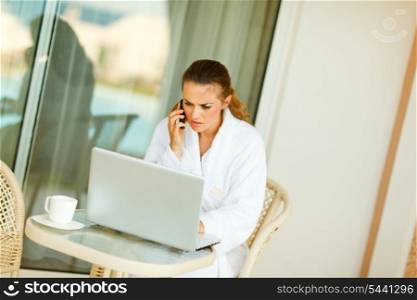 Concerned woman in bathrobe sitting at table on terrace speaking mobile and looking in laptop&#xA;