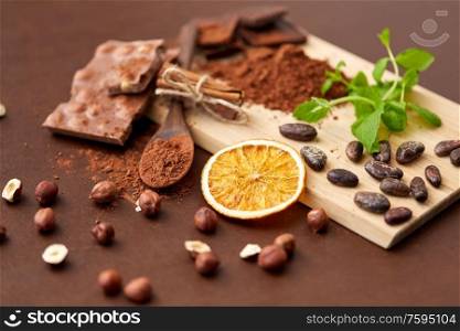 confectionery and culinary concept - chocolate with hazelnuts, cocoa beans, and powder, dry orange and cinnamon on wooden board with spoon on brown background. chocolate with hazelnuts, cocoa beans and orange