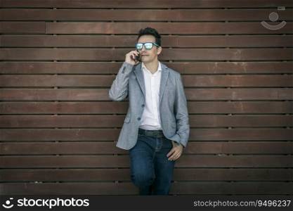 Confident adult businessman dressed in stylish gray jacket and jeans with sunglasses having phone call while standing against plank wooden wall. Elegant man in trendy outfit talking on phone