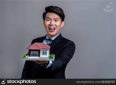 Confident Asian businessman holding house model, advertising home loan with smile. Real estate agent with sample house model in hand on isolated background for housing business advertisement. Jubilant. Confident Asian businessman holding house model. Jubilant