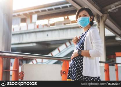Confident Asian Pregnant Woman in face mask looking at camera while traveling to work in the city. Young mother smiling relaxing in public outdoor during pandemic virus. Healthcare with New normal