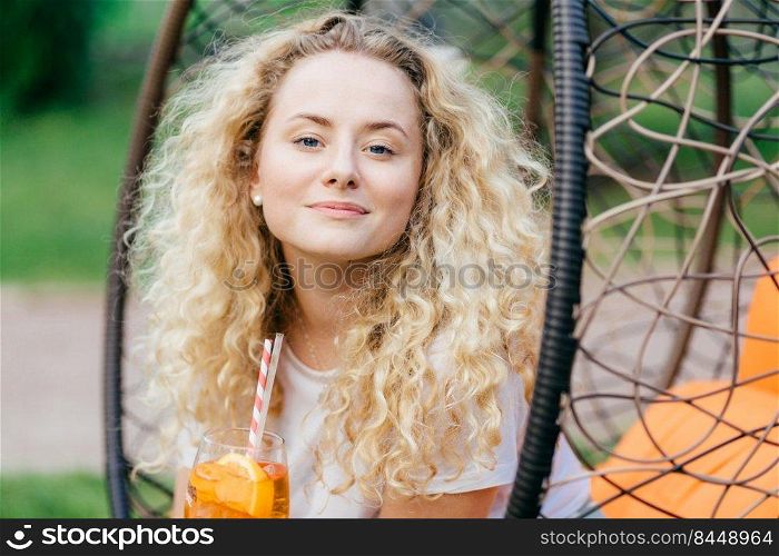 Confident beautiful female with light curly hair, drinks summer cocktail, sits on hanging chair, enjoys good rest alone, poses outdoor. Young woman has healthy skin, rests in garden during sunny day