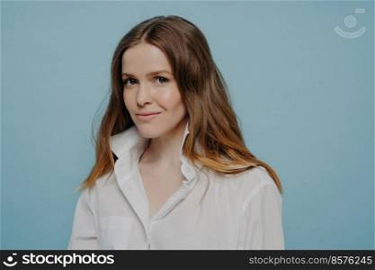 Confident beautiful teenage girl in white shirt with unbuttoned top being assured and looking at camera with slight smile while standing isolated on blue background, studio portrait. Confident beautiful teenage girl in white shirt smiling at camera