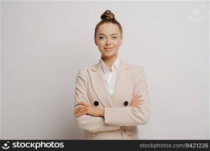 Confident beautiful woman manager in beige suit, white shirt, crossed arms, looking forward, against light wall.