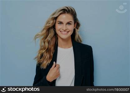 Confident blonde woman dressed in black coat holding collar with one hand and radiating confidence, expressing positive emotion by smiling, isolated next to blue wall. Successful women concept. Blonde businesswoman holding collar with one hand and radiating confidence