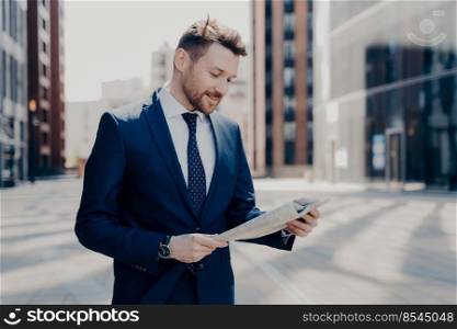 Confident business owner dressed in blue suit feeling delighted after reading positive feedback about his suggested project in local newspaper, walking alone at office. Business owner reading positive news in newspaper while walking to work