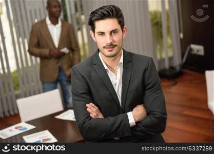 Confident businessman. Cheerful young caucasian man in formalwear keeping arms crossed and smiling while his black colleague standing on background using a smart phone
