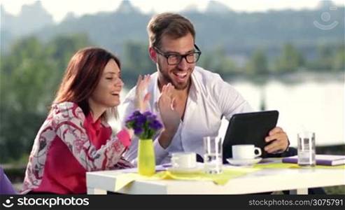 Confident businessman in eyeglasses and his colleague talking with their partner online using tablet during business lunch outdoors