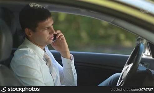 Confident businessman in necktie communicating on smartphone while sitting in car. View from car window. Side view. Handsome entrepreneur discussing business matter with his colleague on mobile phone while sitting in driver&acute;s seat.