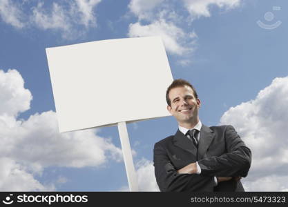 Confident businessman with arms crossed standing by blank sign against cloudy sky
