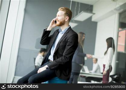Confident businessman with ginger hair using mobile phone while other business people working in office
