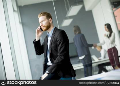 Confident businessman with ginger hair using mobile phone while other business people working in office