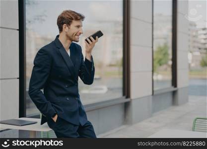 Confident businessman with smartphone using voice assistant app to organize his day or recording message, speaking on speakerphone, standing outside with hand in pocket while leaning on cafe table. Confident businessman with smartphone using voice assistant app while leaning on cafe table outdoors