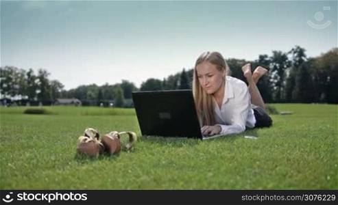 Confident businesswoman lying on green grass with laptop and typing