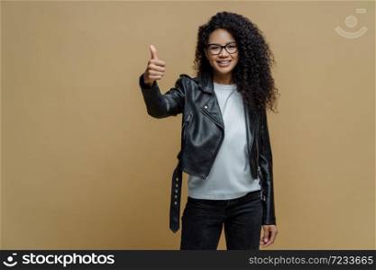Confident cheerful African American woman shows thumb up in approval, gives recommendation or advice dressed in stylish leather jacket smiles positively isolated on beige background. I like and accept
