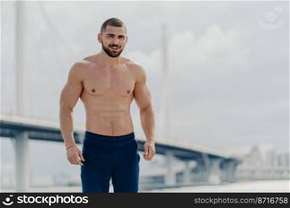 Confident cheerful bearded man with naked torso looks gladfully at camera, dressed in trousers, poses against river bridge, leads healthy lifestyle, has muscular body after regular trainings