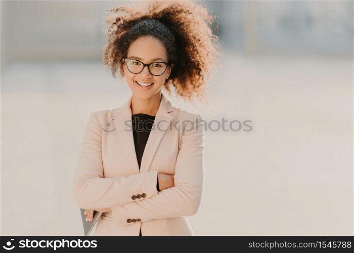 Confident cheerful female entrepreneur with curly Afro hair, keeps arms folded, uses modern tablet computer, smiles positively, waits for business partner outdoor, blurred urban setting in background