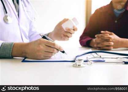 Confident doctor man holding a pill bottle and writing while talking with senior patient and reviewing his medication at office room.