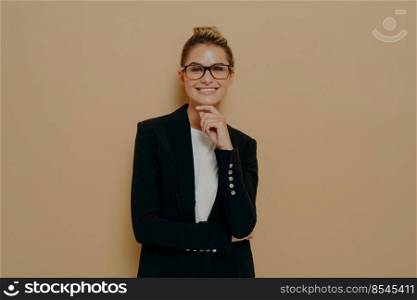 Confident european young female in spectacles and black blazer over white tshirt touching her chin tenderly with hand and smiling widely at camera, expressing positive emotions while posing in studio. Confident european young female in spectacles and black blazer over white tshirt touching her chin tenderly