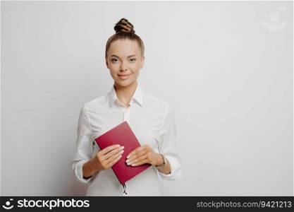 Confident female accountant in white shirt, hair in bun, holds red notebook with company logs and transactions, looks at camera, isolated on grey background.