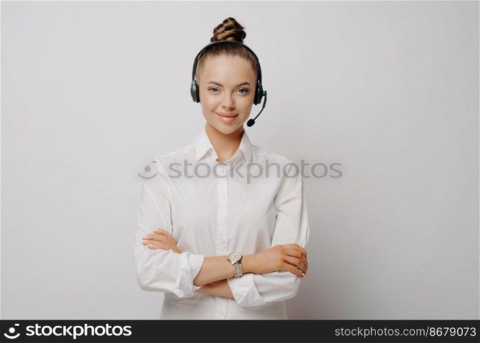 Confident female air traffic controller in shirt wearing black headset, standing with crossed arms communicating with plane pilots, standing against light wall. Portrait of call center worker. Confident female air traffic controller in white shirt
