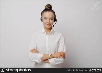 Confident female air traffic controller in shirt wearing black headset, standing with crossed arms communicating with plane pilots, standing against light wall. Portrait of call center worker. Confident female air traffic controller in white shirt