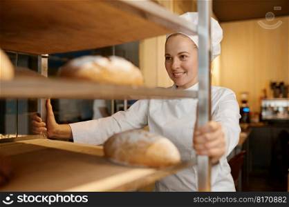 Confident female baker pulling trolley rack of freshly baked bread loaves. Bakeryhouse worker and baking process. Confident female baker pulling rack of freshly baked bread