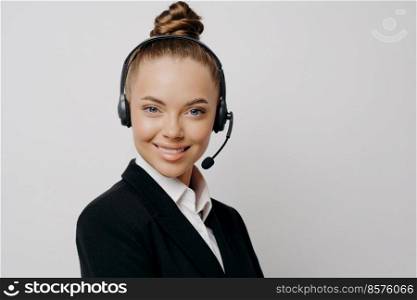 Confident female call center manager in dark suit with black wireless headset looking happily at camera while posing in front of light background. Happy businesswoman having online call in office. Happy businesswoman during online video call in office