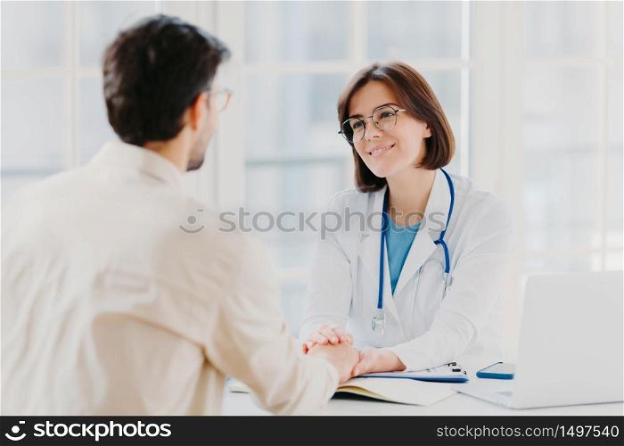 Confident female doctor holds hands of ill patient, persuades everything will be alright, dressed in white medical gown, gives advice, pose in hospial office. Consultation and diagnosis concept