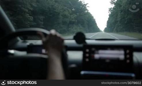 Confident female driver driving car on rural road through the forest. View from inside out of vehicle&acute;s interior. Woman travelling by car on asphalt road in countryside during summer road trip.