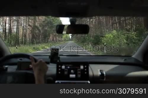 Confident female driver driving car through the forest on two lane rural road on summer day. View form inside of vehicle. Asphalt country road throgh the woods.