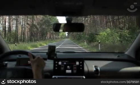 Confident female driver driving car through the forest on two lane rural road on summer day. View form inside of vehicle. Asphalt country road throgh the woods.