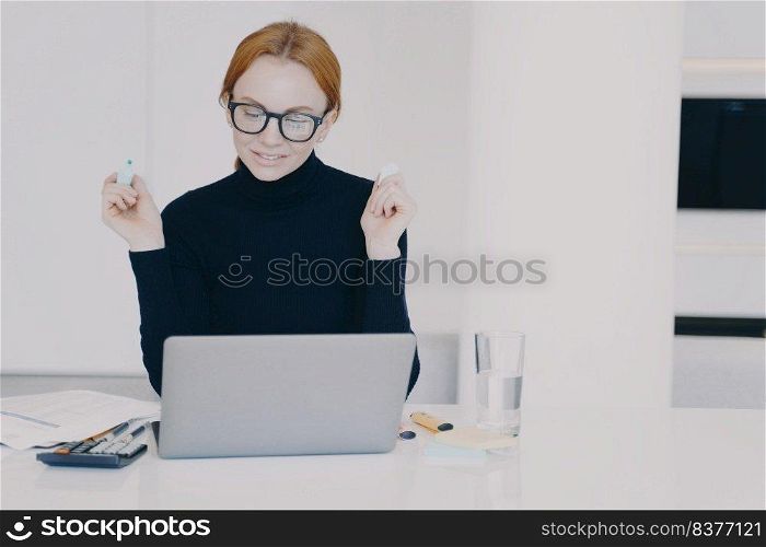 Confident female entrepreneur at work. Red haired woman in office is doing paperwork with marker pen and highlighting text. Positive caucasian girl entrepreneur is working on project at laptop.. Confident female entrepreneur woman in office is doing paperwork with marker pen highlighting text.