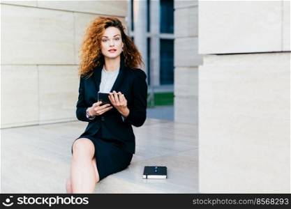 Confident female freelancer being busy while working with her tablet, writing notes in her pocket book, wearing black formal costume, looking directly into camera. Office worker using modern gadget