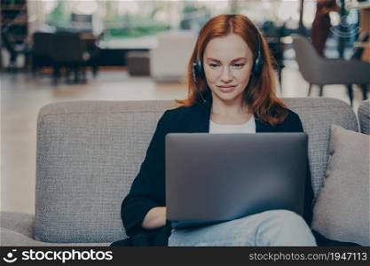 Confident focused freelancer redhead woman using headset earphones and laptop, working remotely online, browsing internet, communicates with customers while sitting on sofa in public place. Confident smiling businesswoman using headset earphones and laptop, working remotely online