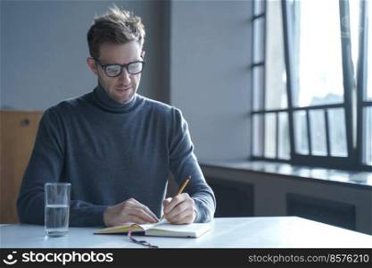 Confident focused young German man freelancer in glasses taking notes in agenda while sitting at desk at home interior, has break from work online, writing down some new fresh ideas that come to mind. Confident focused German man freelancer in glasses taking notes in agenda sitting at desk at home