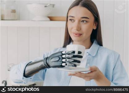 Confident handicapped girl is holding cup of tea with cyber hand. Happy disabled girl at kitchen at home. Pretty woman with makeup is smiling. Concept of grasp sensors in modern electronic prosthesis.. Confident handicapped girl holding cup of tea with cyber hand. Happy woman with makeup is smiling.