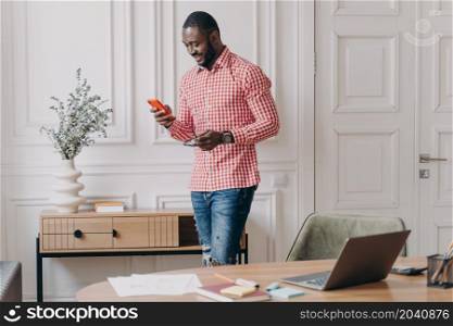 Confident handsome african american businessman using mobile, standing in office during working day, young dark skinned man employee typing and looking at phone screen with smile, chatting online. Confident handsome african american businessman using mobile, standing in office during working day