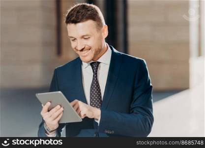 Confident handsome male manager checks email on tablet computer, updates database on touch pad, wears elegant formal clothing, connected to wireless internet, poses outdoor. Business concept