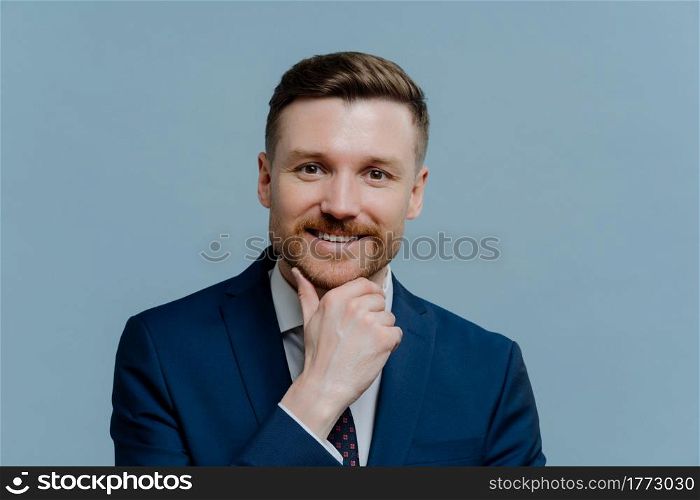 Confident happy businessman in formal wear touching his face with stubble, looking happily at camera while standing isolated over light steel blue background, positive male employee feeling satisfied. Headshot of successful happy businessman in suit looking at camera