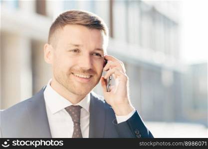 Confident happy middle aged businessman communicates via cell phone, discusses financial report with business partner, wears formal clothes, stands outdoor against blurred background. Busy CEO