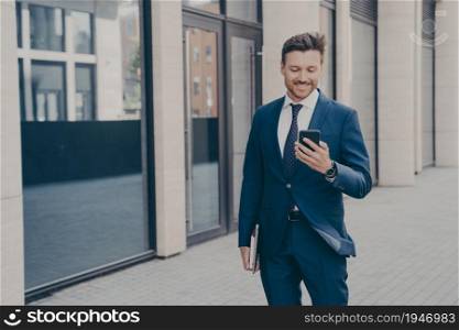 Confident happy young male business consultant holding modern smartphone and smiling while standing outdoors after working day, reading sms from college with good news about project. Confident happy young male business consultant holding modern smartphone and smiling outdoors