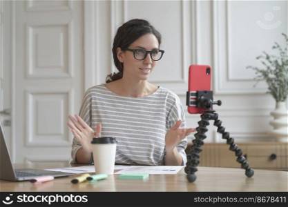 Confident Italian woman economist counselor recording video stream online consultation for group and individuals, tutor lady using phone on tripod for video chat sitting at home. Business blog concept. Confident Italian woman economist counselor recording video stream online using smartphone