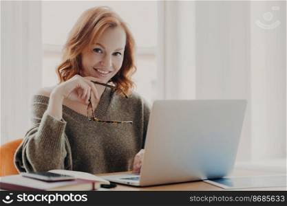 Confident joyful redhead woman works on modern laptop, poses in coworking space, wears sweater, works remotely with application on device, smiles pleasantly, holds spectacles, enjoys distance job