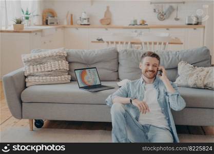 Confident male freelance worker calling his employer to tell him good news about project, making another client satisfied, laptop with infographics, sitting on floor while resting against couch. Confident male freelance worker calling his employer to tell him good news about project