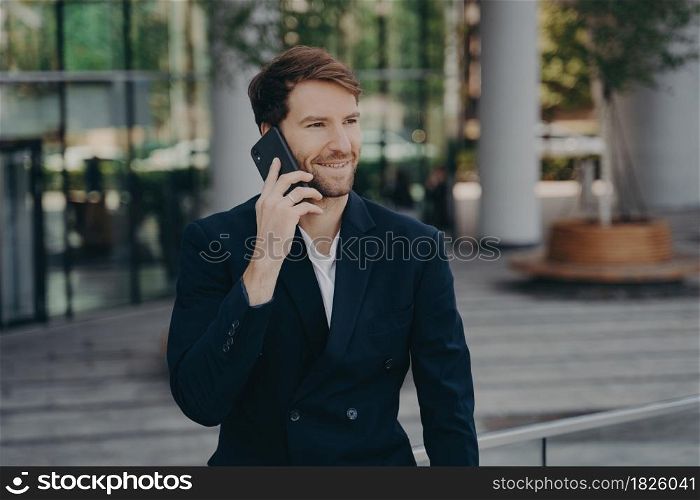 Confident man entrepreneur dressed in formal clothes makes phone call in roaming looks away has cheerful expression poses near office building enjoys telephone conversation during work break. Confident man entrepreneur dressed in formal clothes makes phone call in roaming